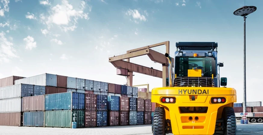 Empilhadeira pesada Hyundai operando em pátio de contêineres, com pilhas de containers ao fundo e guindaste portuário.