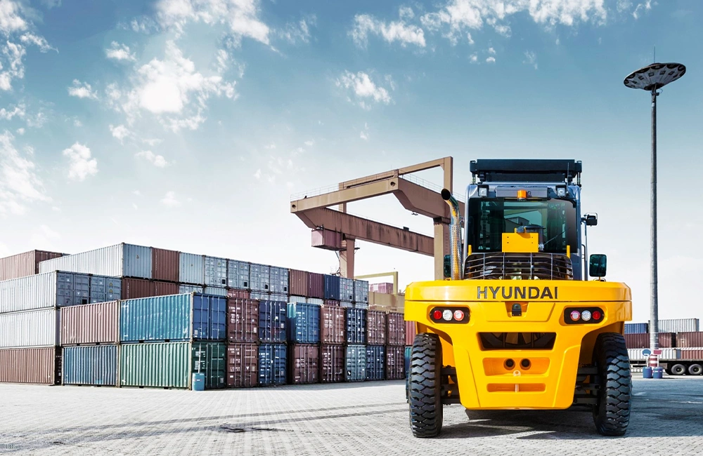 Empilhadeira pesada Hyundai operando em pátio de contêineres, com pilhas de containers ao fundo e guindaste portuário.