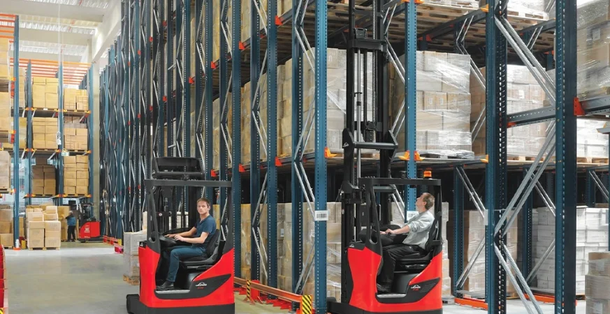 Operadores utilizando empilhadeiras retráteis em um armazém de estocagem alta, movimentando pallets em estrutura porta-pallets vertical.