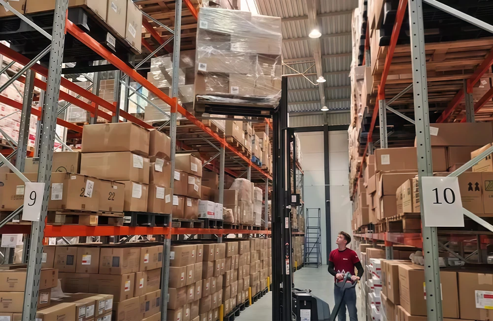 Operador utilizando empilhadeira retrátil em corredor alto de armazém organizado com pallets e caixas.