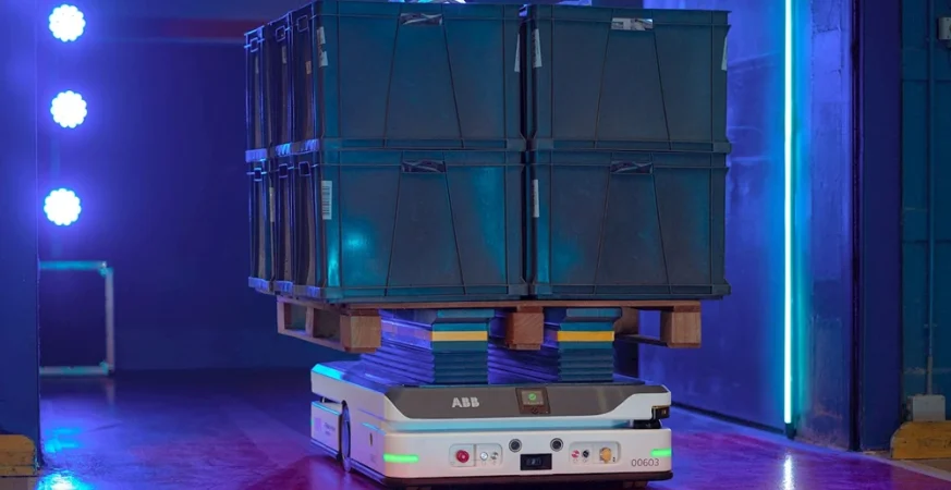 Robô móvel autônomo transportando caixas empilhadas sobre palete em ambiente industrial iluminado.