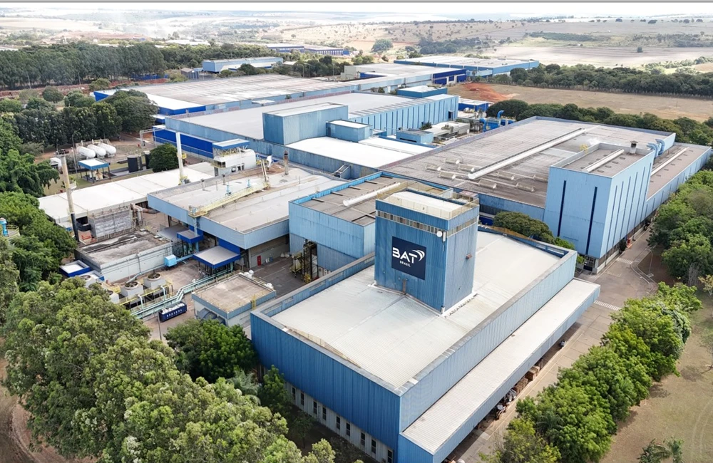 Vista aérea da planta industrial da BAT Brasil, com galpões e áreas operacionais cercados por vegetação, representando iniciativas de redução de emissões e eficiência ambiental.