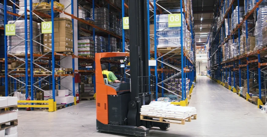 Empilhadeira elétrica operando em galpão logístico, movimentando pallet entre estruturas porta-pallets altas em ambiente de intralogística.