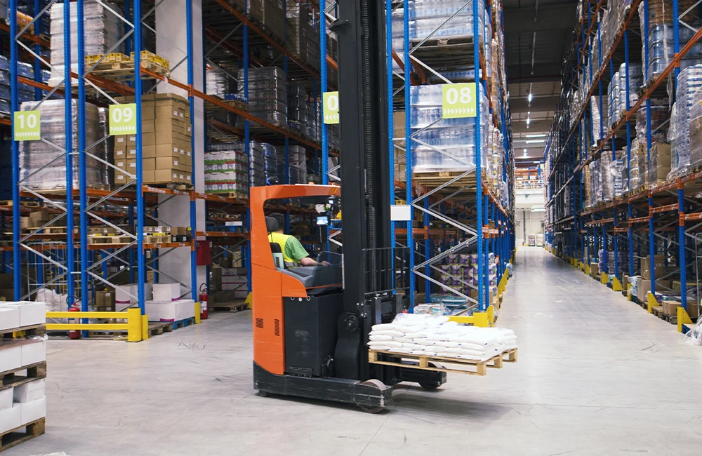 Empilhadeira elétrica operando em galpão logístico, movimentando pallet entre estruturas porta-pallets altas em ambiente de intralogística.