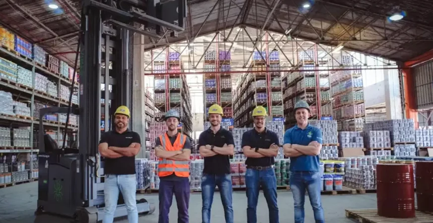 Equipe em armazém industrial ao lado de empilhadeira de torre com corredores altos e produtos estocados.