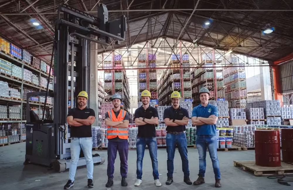Equipe em armazém industrial ao lado de empilhadeira de torre com corredores altos e produtos estocados.