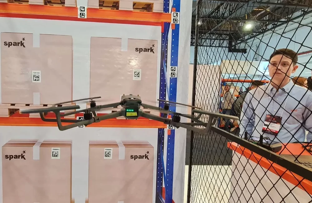 Drone realizando leitura de códigos em estante de armazenagem durante demonstração de inventário logístico automatizado.