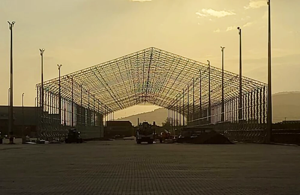 Estrutura metálica de um galpão lonado em construção ao entardecer, com colunas e treliças montadas em área logística.