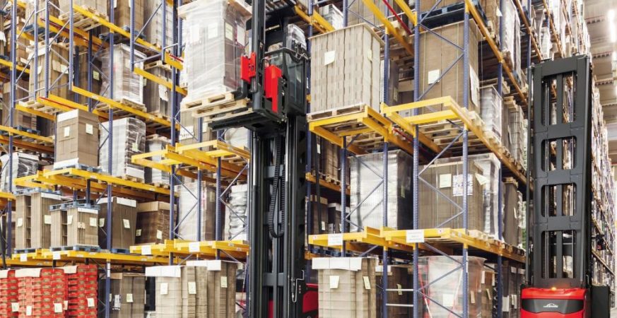 Empilhadeira retrátil operando entre corredores altos de armazenagem, movimentando paletes em estantes verticais.