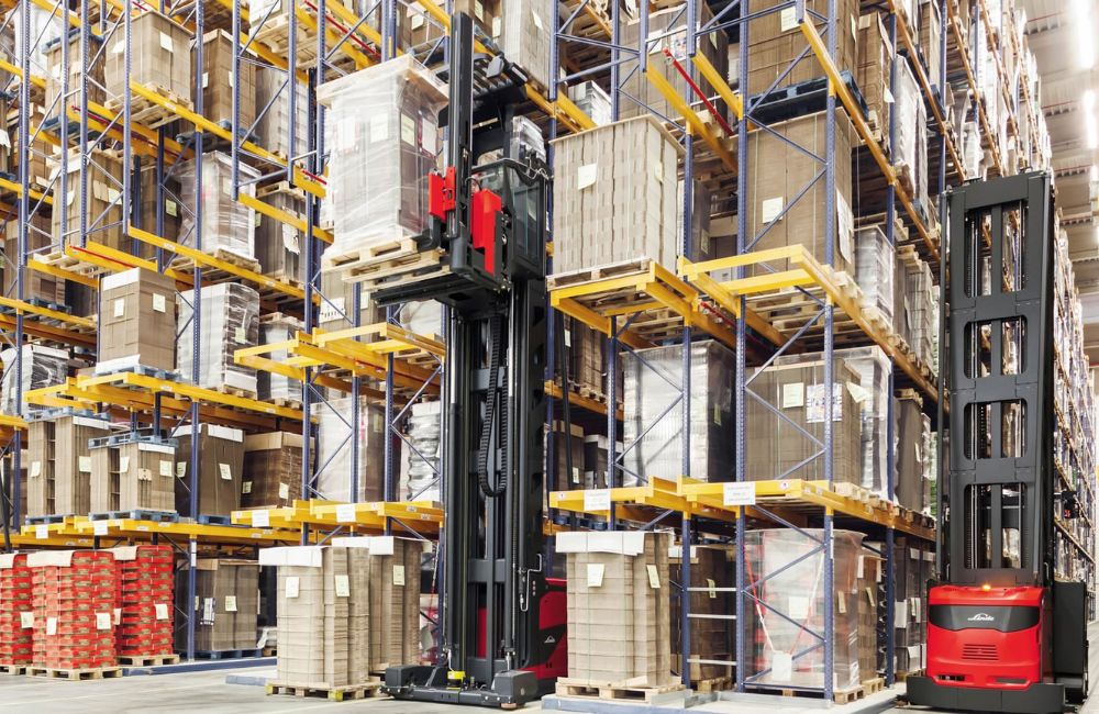 Empilhadeira retrátil operando entre corredores altos de armazenagem, movimentando paletes em estantes verticais.