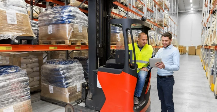 Operador utilizando dispositivo de telemetria ao lado de empilhadeira em operação dentro de centro de distribuição.