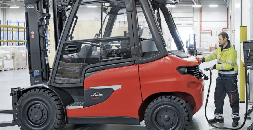 Empilhadeira elétrica conectada ao carregador em armazém logístico, ilustrando a gestão de energia para prolongar a vida útil da bateria e do equipamento.